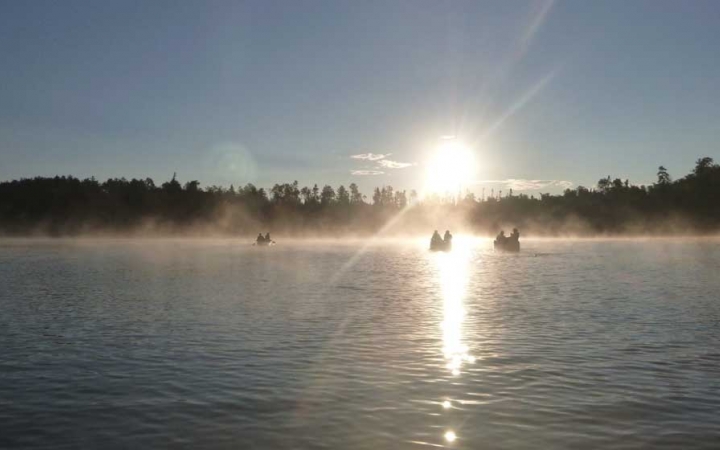 In the distance, three canoes are paddled toward the sun, through fog sitting on the water. 